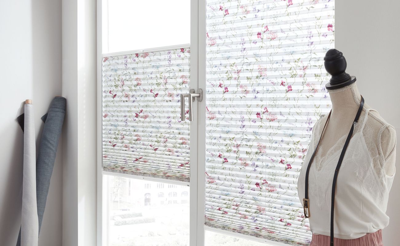 Fenster mit dekorativen Jalousien in Blumenmuster und eine Schneiderpuppe in einem Raum.