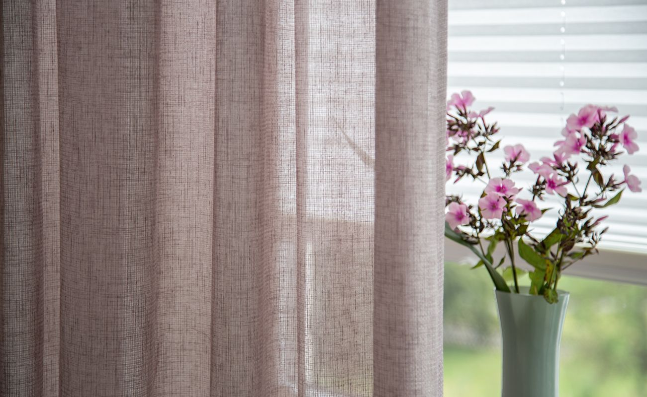 Ein heller Vorhang aus transparentem Stoff mit einem Blumenstrauß in einer Vase auf einem Fensterbrett.